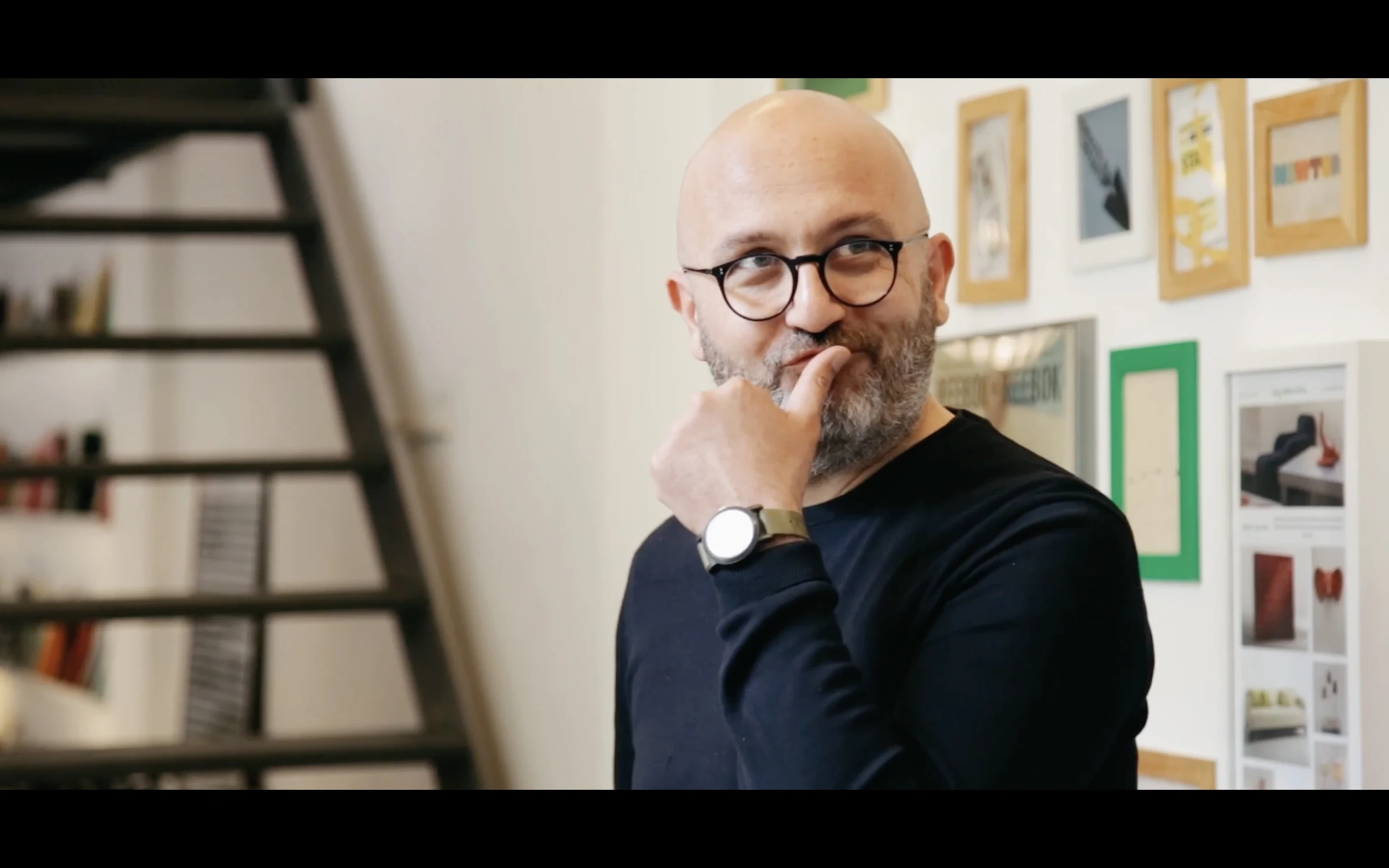 A designer in glasses pausing mid-thought, framed by artwork on the wall