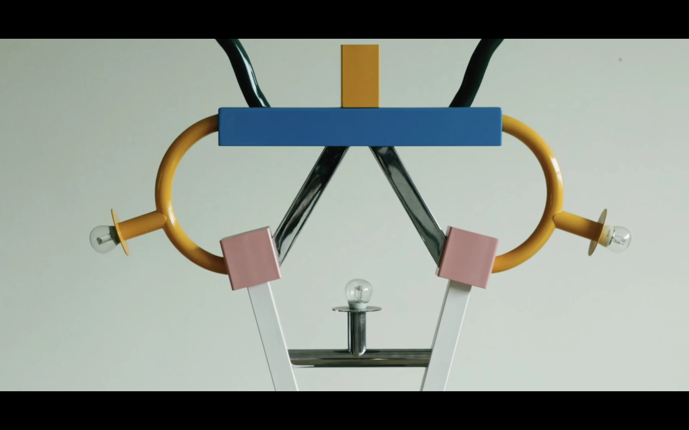 A sculptural object in yellow, blue, and pink balanced on slender legs